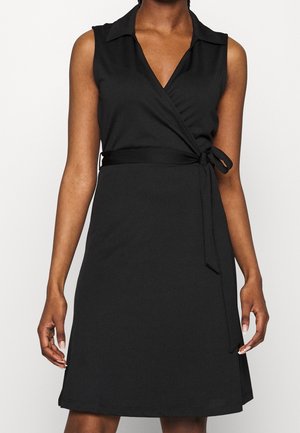Robe portefeuille noire sans manches avec col et ceinture nouée sur le côté, portée par une personne sur un fond uni.