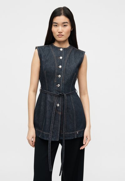 Νεαρή γυναίκα που φοράει αμάνικο σκούρο τζιν τοπ με κουμπιά και ζώνη στη μέση, στέκεται μπροστά σε απλό λευκό φόντο.