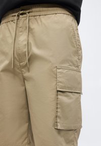 Μπεζ κοντά cargo παντελόνια με ελαστική μέση και κορδόνι, με μεγάλη τσέπη με καπάκι στο δεξί μηρό, σε απλό φόντο.