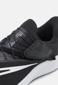 Chaussure de sport noire avec une tige texturée, un accent Swoosh blanc, et la technologie FlyEase. Elle dispose de lacets élastiques et d'une semelle flexible.