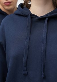 Tamsiai mėlynas džemperis su gobtuvu, pagamintas iš minkštos medžiagos, turi raištelį prie gobtuvo ir tekstūruotus akcentus. Artimas vaizdas pabrėžia medžiagos lygų paviršių.