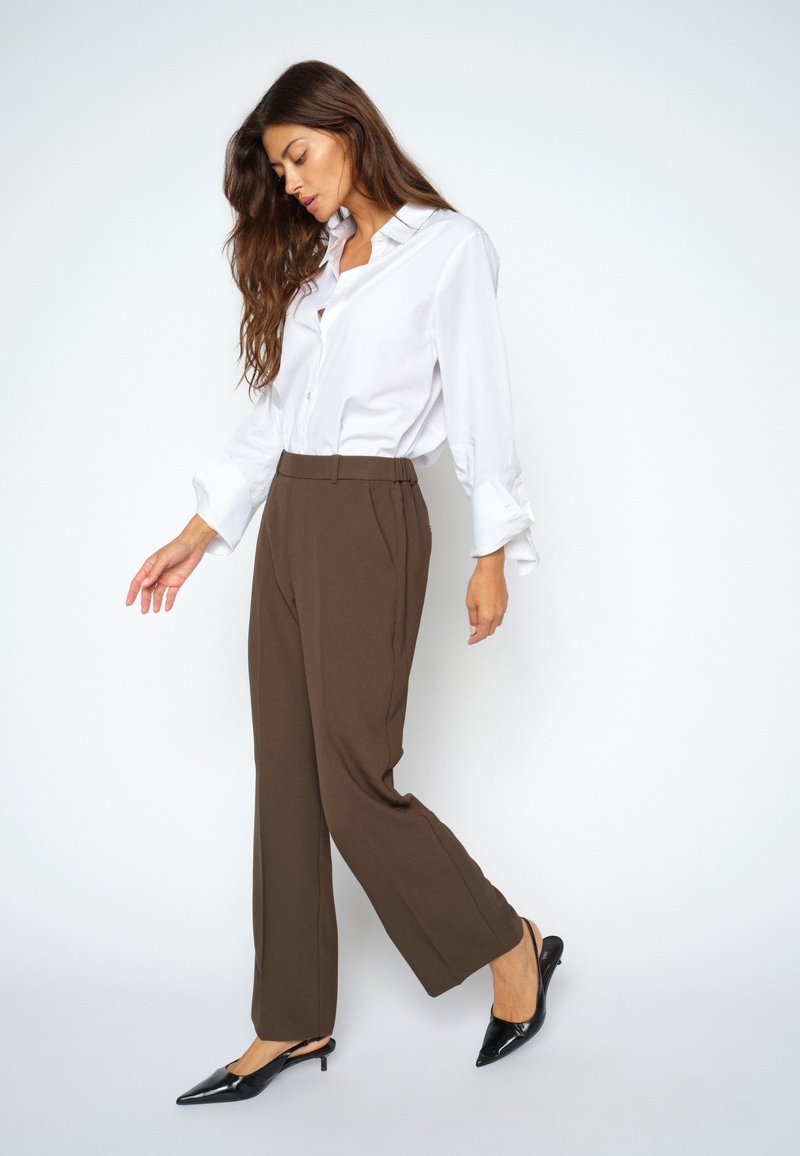 Donna con lunghi capelli castani che indossa una camicia bianca con bottoni, pantaloni marroni a gamba larga e scarpe nere con tacco basso che cammina su uno sfondo bianco.