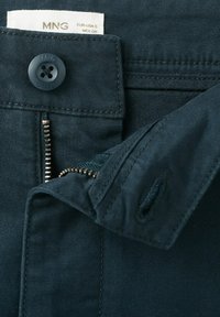 Pantalon bleu foncé avec une fermeture éclair ouverte, un bouton marqué "MNG" et une étiquette de taille visible à l'intérieur de la ceinture.