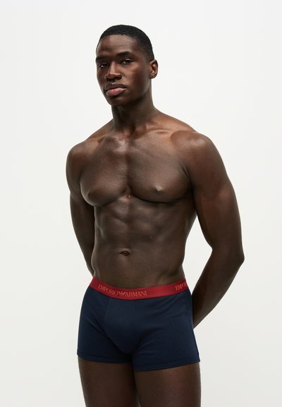 Homme musclé à la peau foncée, debout, mains dans le dos, portant un boxer Emporio Armani bleu marine avec une ceinture rouge.