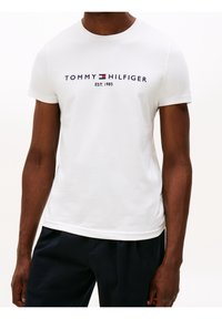 Camiseta blanca de algodón con mangas cortas, con la inscripción "TOMMY HILFIGER EST. 1985" en letras azul marino y rojas en el pecho. Textura plana.