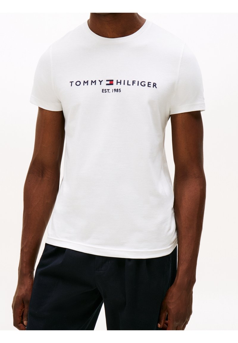 Camiseta blanca de algodón con mangas cortas, con la inscripción "TOMMY HILFIGER EST. 1985" en letras azul marino y rojas en el pecho. Textura plana.
