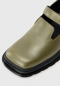 Zeleno usnjeno čevlje z bleščečo površino, okroglo konico in črnim gumijastim podplatom. Ima trak čez vrh za varno prileganje.