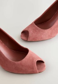 Offene pinke Wildlederabsätze mit glatter Oberfläche, abgerundetem Design und tief ausgeschnittenen Seiten. Die Schuhe haben eine konturierte Form und eine weiche Textur.