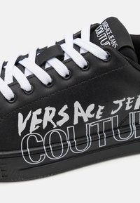 Czarne sneakersy z białymi sznurówkami, z wyraźnym czarno-białym brandingiem. Gładki skórzany materiał i teksturowana gumowa podeszwa.