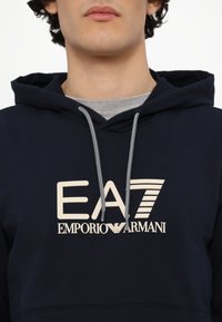 Navy hoodie made of cotton featuring a large ivory EA7 Emporio Armani logo, drawstring hood, and ribbed cuffs. Simple, modern design.