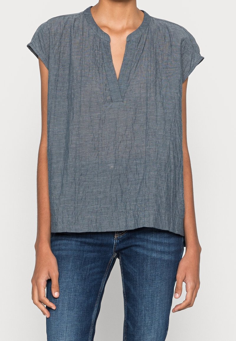 Femme portant un chemisier gris froissé à manches courtes avec un col en V, assorti à un jean en denim bleu sur un fond uni.