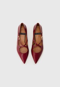 Burgundy leather high heels with a pointed toe, cut-out design, and crossed straps featuring a small gold buckle accent. Smooth texture.
