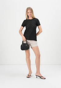 Black short-sleeve top with cut-out patterns, paired with beige shorts. Holding a small black textured handbag, wearing black flip-flops.