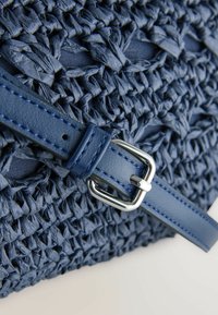 Gros plan sur un sac tissé bleu marine avec une sangle en cuir lisse et une boucle en argent brillant sur un fond clair.