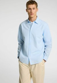 Camisa abotonada azul claro con un patrón texturizado, con cuello extendido y mangas largas, combinada con pantalones beige.