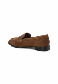 Mocasines de ante marrón con un bajo tacón negro, que presentan una puntera cosida y un acento metálico decorativo en la parte superior. Textura suave, forma clásica.