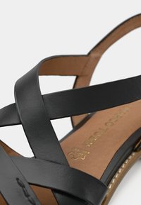 Μαύρες δερμάτινες σανδάλια με σταυρωτές ζώνες, καφέ μαλακή σόλα και λεπτομερή ραφή κατά μήκος των άκρων.