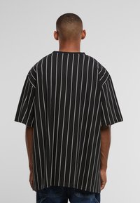 Mann, der mit dem Rücken zur Kamera steht, trägt ein schwarzes übergroßes T-Shirt mit vertikalen weißen Nadelstreifen und dunkle Jeanshose vor grauem Hintergrund.