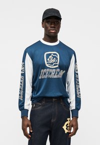 Μπλε μακριμάνικο πουλόβερ από μεσάντζ με λευκά μανίκια, με γραφικό λογότυπο και κείμενο στο μπροστινό μέρος και στα μανίκια. Σκούρο τζιν παντελόνι με μπαλώματα.