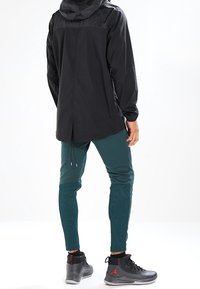 Men's black hooded jacket with a matte finish, paired with dark green tapered pants featuring textured accents, and grey sneakers with red details.