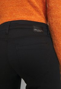 Jeans de mezclilla negra con una textura suave, que cuentan con bolsillos traseros y una etiqueta de Mavi Jeans Co. El suéter naranja que lo acompaña tiene un aspecto suave y peludo.