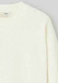 Pull en maille écru avec une texture côtelée, un col rond et un design à épaules tombantes. L'étiquette indique la taille "S" de la marque BZB.