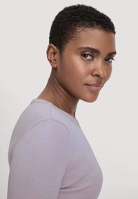 Lichtpaarse getailleerde longsleeve shirt met een ronde hals, een gladde textuur en minimale stikdetails. Model toont het shirt vanuit een zijaanzicht.