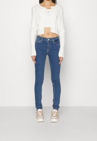 Cárdigan blanco corto, jeans ajustados azules, zapatillas beige. Textura de punto suave, estilo clásico de mezclilla, detalle sutil del logo en los jeans.