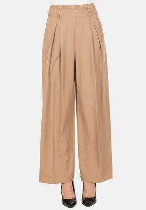 Pantaloni beige a gamba larga con pieghe, vita alta e vita elastica indossati con tacchi a punta nera su uno sfondo bianco.
