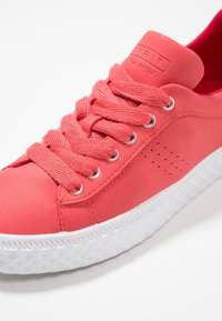 Baskets rose corail avec lacets épais, détails perforés sur le côté, semelle blanche texturée et logo en relief sur la languette rembourrée.