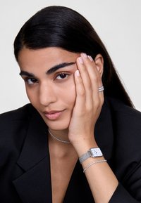 Montre carrée argentée avec bracelet en maille, associée à un délicat bracelet en argent et un choker, portée sur une main près d'un visage, blazer noir.