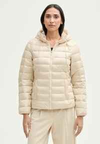 Veste matelassée beige avec capuche, fermeture éclair à l'avant et poches zippées. Présente un design matelassé avec des coutures horizontales et des poignets élastiques.