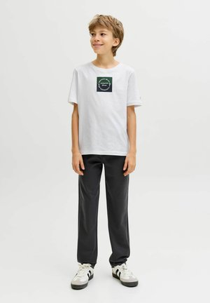 Witte katoenen T-shirt met een vierkante groene afbeelding op de borst, gecombineerd met zwarte, loszittende broek en grijze sneakers met zwarte strepen.