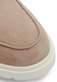 Zapato de ante beige con punta redonda, suela de goma blanca y costuras en contraste a lo largo del borde superior, que resaltan la textura y suavidad.