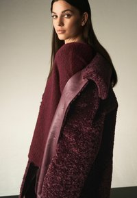 Bordeaux sweater med et struktureret strik, parret med en plysset yderjakke med læderdetaljer og et draperet design.
