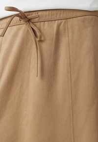 Jupe en daim marron clair en gros plan montrant une ceinture nouée et un détail de couture verticale sur un tissu lisse.