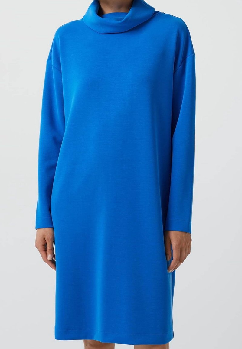Femme portant une robe bleu vif à manches longues et longueur genoux avec un col roulé ample sur un fond clair uni.