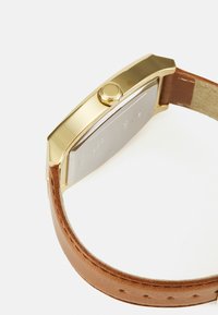 Relógio de pulso em tom de ouro com caixa retangular, pulseira de couro castanho e detalhe polido no botão. Fundo prateado com texto gravado.