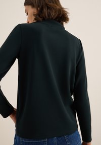 Dunkelgrüner Pullover mit langen Ärmeln, aus weichem Stoff gefertigt. Hat einen hohen Kragen und eine glatte Textur. Der Saum ist gerade und leicht verlängert.