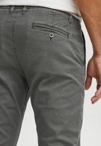 Pantaloni grigi in misto cotone con una texture liscia, dotati di una tasca posteriore con chiusura a bottone e una leggera lucentezza.