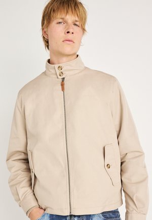 Giacca marrone chiaro con colletto alto, chiusura frontale con zip e due tasche laterali. Presenta dettagli marrone chiaro e un tessuto liscio e strutturato.