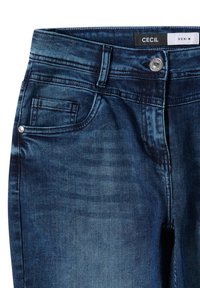 Granatowe dżinsy z przodu z kieszeniami, szlufkami na pasek, srebrnym guzikiem i metką z napisem "CECIL DENIM" na pasku.