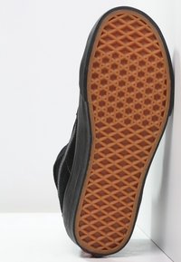 Semelle d'une chaussure noire avec une semelle en caoutchouc marron à motif gaufré, positionnée verticalement contre un fond blanc et un coin de mur.