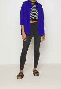 Camisa azul de gran tamaño sobre un suéter recortado de patrón en blanco y negro, combinada con jeans negros ajustados y sandalias negras con hebillas.