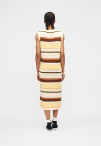 Άτομο με πλεξούδες, φορώντας αμάνικο φόρεμα μέσης μήκους με ρίγες σε γήινους τόνους και μαύρα ψηλά αθλητικά παπούτσια, στέκεται γυρισμένο με την πλάτη στην κάμερα.