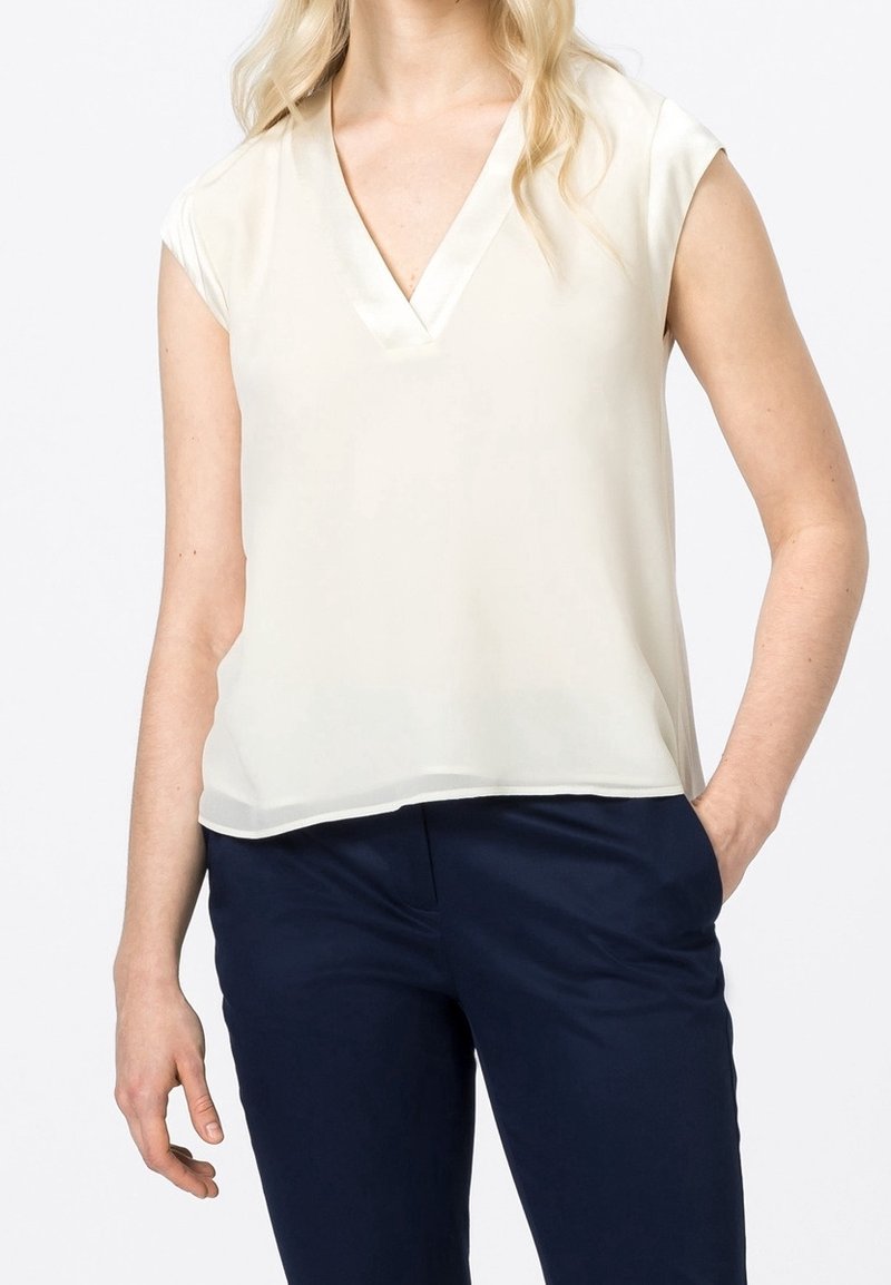 Blouse crème sans manches avec un décolleté en V profond et bordure en satin. Le tissu est lisse et léger, assorti à un pantalon bleu marine.