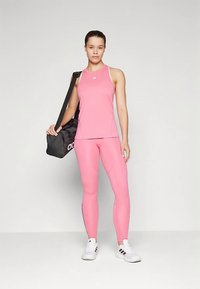 Žena v růžovém sportovním topu a legínách stojí a drží černou tašku Adidas přes rameno, na nohou má bílé tenisky Adidas.