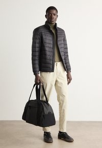 Zwarte pufferjack, olijfgroene coltrui, lichtbeige broek. Zwarte en grijze sporttas met twee hengsels. Donkere schoenen, minimalistisch ontwerp.