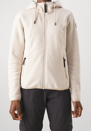 Person trägt einen cremefarbenen, strukturierten Kapuzenpullover mit Reißverschluss und Kordeln sowie schwarze Shorts, steht vor einem schlichten Hintergrund.