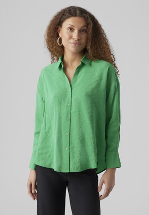 Chemise verte à col ouvert en tissu léger, avec une coupe ample et des manches longues. Portée avec un pantalon noir.